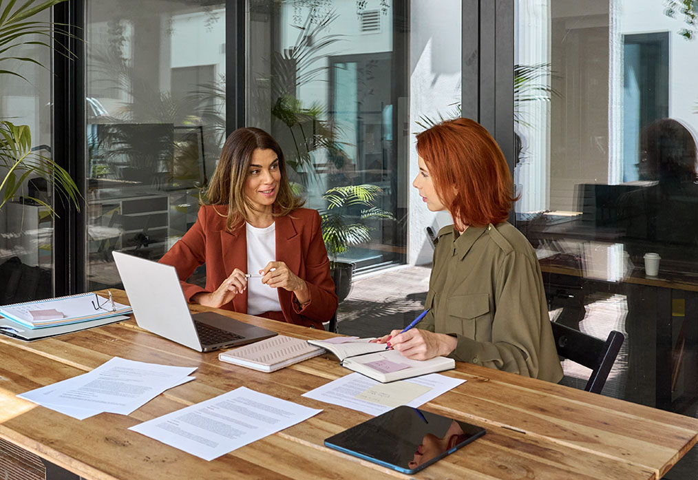 Two women engaged in a business discussion at a wooden table, with a laptop and documents, emphasizing collaboration and tailored marketing strategies for small to medium-sized businesses.
